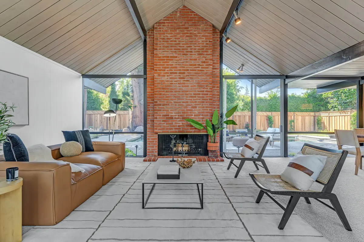 Living room at 1674 Alexander Way: a full-height red brick chimney between two glass walls, under a gable of exposed beams and painted tongue-and-groove decking.