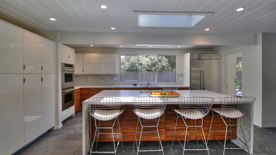 Kitchen at 1694 Clay Drive: white gloss and walnut cabinetry, a large stone-topped island with wire stools, and a skylight cut into the flat roof above it.
