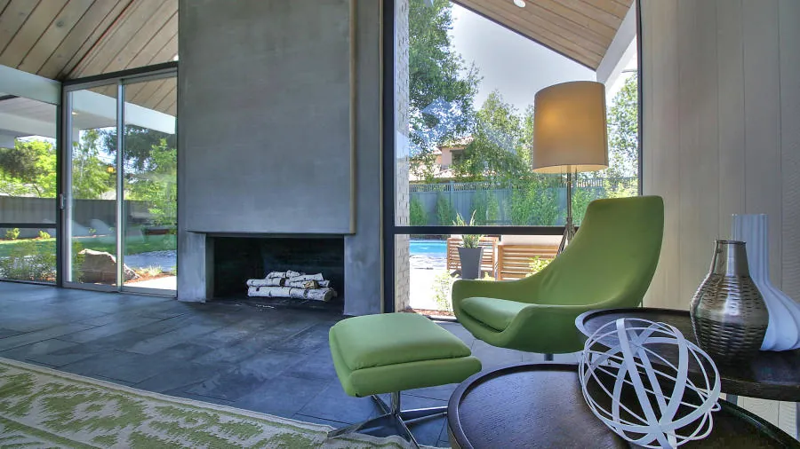 Living room at 1694 Clay Drive: a concrete fireplace mass between two glass walls meeting at an open corner, under a wood gable ceiling, with the pool beyond.